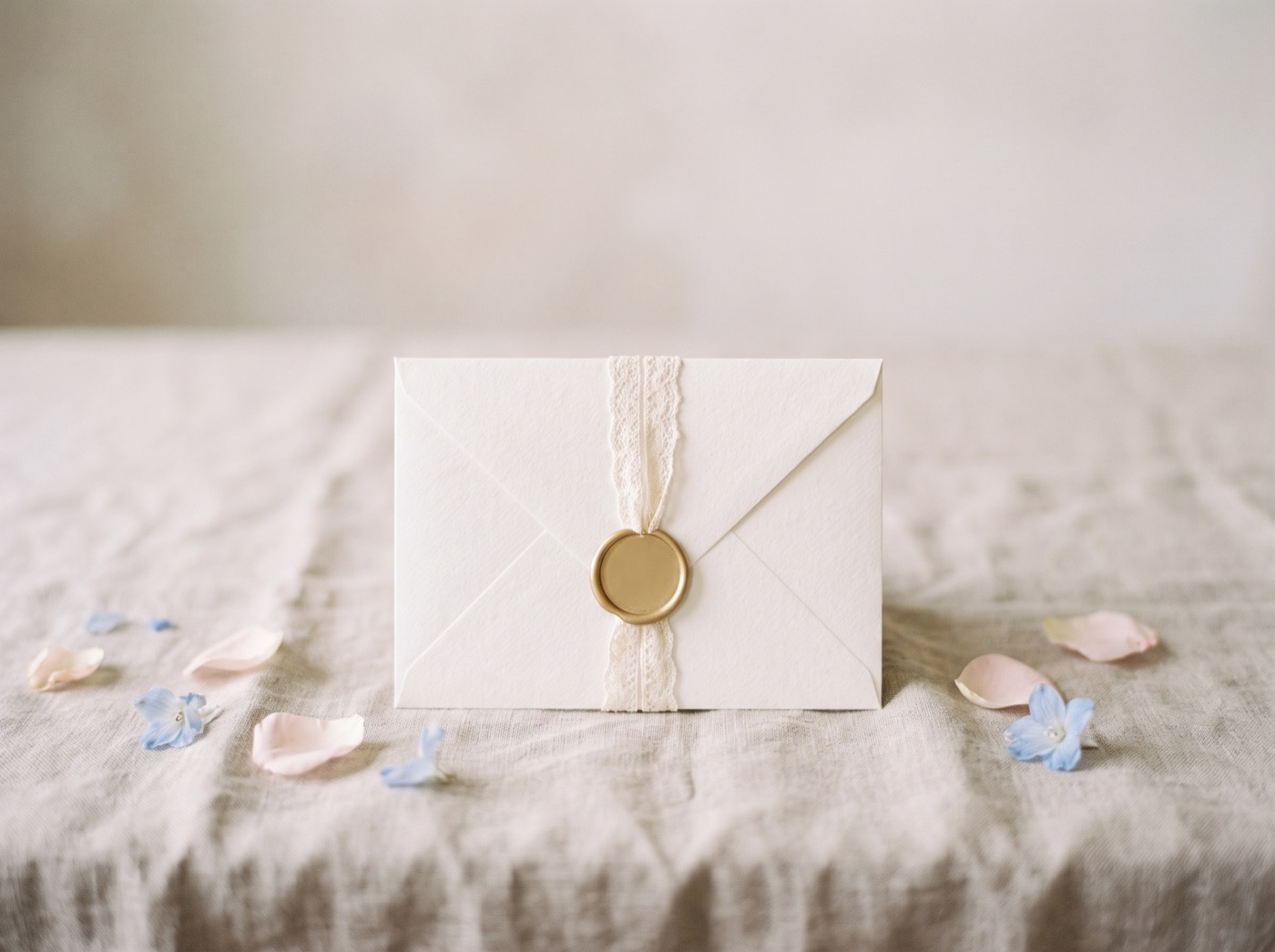 An ivory envelope with a lace band and a gold wax seal, with petals scattered on linen